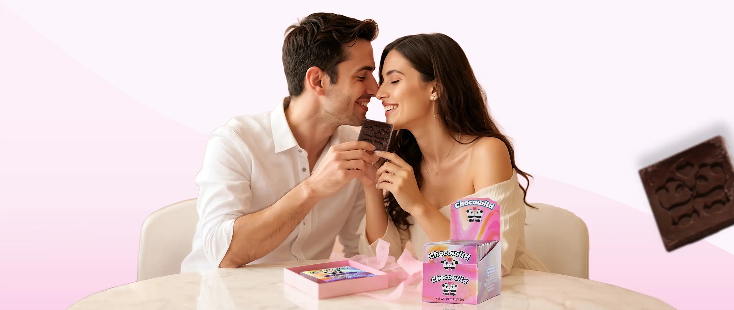 Smiling couple sitting at a table, sharing a piece of Chocowild chocolate with pastel-colored packaging in the foreground, set against a soft pink background.